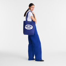 Load image into Gallery viewer, person holding Longchamp Le Pliage Constantin Riant Collection Medium Tote Bag in color Cobalt blue with Lonchamp Paris 1948 graphic in white