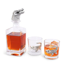Load image into Gallery viewer, Square glass decanter with silver colored alligator head bottle stopper and two short glasses with silver colored alligator motifs on the side of the glass decanter and one glass filled with amber liquid