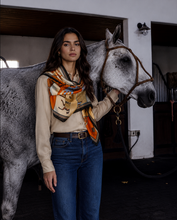 Load image into Gallery viewer, A woman stands indoors beside a gray horse, wearing a cream long-sleeve top, blue jeans, and a patterned silk scarf with orange, black, and beige tones. The horse has a brown halter.