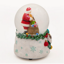 Load image into Gallery viewer, A festive snow globe figurine featuring Santa Claus packing a Christmas sack, set inside a clear glass globe on a speckled white base decorated with snowflakes and a green garland.