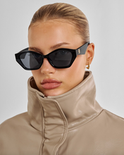 Load image into Gallery viewer, A close-up of a model wearing black cat-eye sunglasses with dark lenses and a beige high-collar jacket against a light gray background.