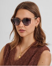 Load image into Gallery viewer, A woman with brown hair is wearing cat-eye sunglasses with a tortoiseshell pattern and gold metal frames, paired with a brown knit top and gold jewelry against a light gray background.