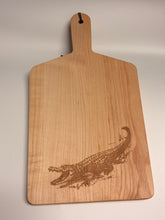 Load image into Gallery viewer, A rectangular wooden cutting board with a handle, featuring a laser-engraved alligator design near the bottom right. The board is made of light-colored wood with visible grain and has a small hole in the top of the handle for hanging.