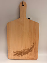 Load image into Gallery viewer, A rectangular wooden cutting board with a handle, featuring a dark brown engraved alligator design on the right side and a black hanging loop on the left, shown against a plain light background.