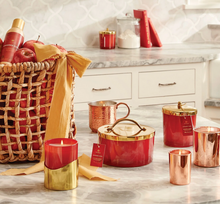 Load image into Gallery viewer, A styled collection of home fragrance and decor items on a marble countertop in a kitchen setting, including red and copper candles, a copper mug, a red and gold lidded container, a red bottle with a gold cap, and a woven basket filled with red apples and a gold spray bottle.