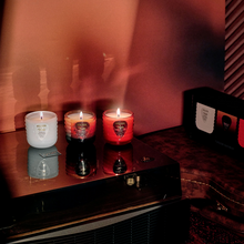 Load image into Gallery viewer, Three lit Kala candles in glass jars are arranged on a glossy black surface in a dimly lit room. The candles come in white, dark amber, and red-orange glass containers, with a branded box visible in the background.