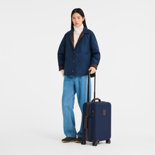 Load image into Gallery viewer, person standing next to the Longchamp Boxford Cabin Suitcase - Small - Embossed Design in Navy Blue - case comes to slightly above her knees

