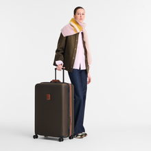 Load image into Gallery viewer, A person standing next to the Longchamp Boxford Suitcase Embossed Style xl brown - suitcase reaches her mid thighs 

