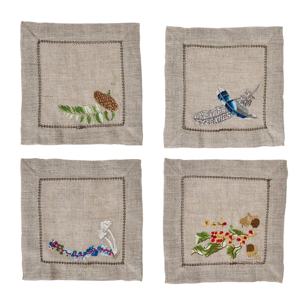 four tan napkins each with a different embroidered design, a pine cone, mushroom and berries, feathers, and leaves, berry and acorns 