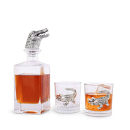 Square glass decanter with silver colored  alligator head bottle stopper and two short glasses with silver colored alligator motifs on the side of the glass