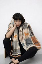 Load image into Gallery viewer, A woman sits on the floor against a light gray wall, wrapped in a beige patchwork scarf with horse illustrations and black leggings.
