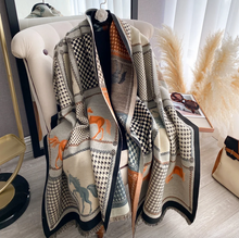 Load image into Gallery viewer, A patterned knit throw blanket draped over a cream tufted armchair in a styled interior. The blanket features a patchwork design with black-and-white checkered and dotted sections, tan and rust-orange horse motifs, and black fringe.
