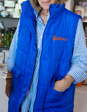 Load image into Gallery viewer, A person wearing a bright blue quilted puffer vest over a blue and white striped button-down shirt. The vest has 'Gators' embroidered in orange on the left chest.
