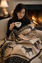 Load image into Gallery viewer, A woman sits on a beige armchair in a cozy living room, wrapped in a patterned throw blanket while reading a book and holding a white mug. The blanket features a cream and black design with geometric motifs and horse silhouettes.
