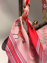 Load image into Gallery viewer, Pink and red striped scarf with black illustrations on a gold chain, attached to a pink textured bag.
