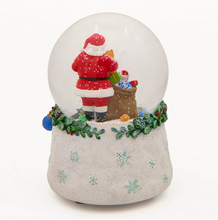 Load image into Gallery viewer, A festive snow globe figurine featuring Santa Claus in a red suit standing next to a brown gift sack filled with presents, set in a clear glass dome on a textured light gray base decorated with snowflakes and green garland.
