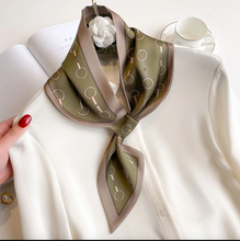 Load image into Gallery viewer, A silk scarf with a horse-bit pattern in olive green and taupe, styled on a white blouse next to an open book and a teacup.
