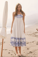 person on the beach wearing a midi length white dress with blue embroidery at the bust and hem