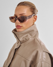 Load image into Gallery viewer, A close-up side profile of a model wearing cat-eye sunglasses with dark brown lenses and a beige high-collar jacket against a light gray studio background.
