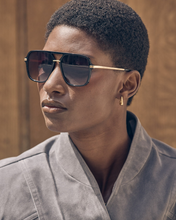 Load image into Gallery viewer, A close-up portrait of a model wearing square aviator-style sunglasses with a black frame, dark lenses, and gold metal temples, paired with a gray collared shirt against a warm brown wooden background.
