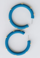 Load image into Gallery viewer, A pair of chunky, open hoop earrings in a vibrant blue color, decorated with small gold studs along the outer curve, displayed on a clean white background.
