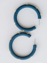 Load image into Gallery viewer, A pair of chunky, open hoop earrings in a matte dark teal blue, embellished with small gold studs along the outer edge, displayed on a clean white background.
