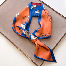 Load image into Gallery viewer, A square silk scarf with a vibrant orange base, featuring a blue border and a floral pattern in white, gray, and black, styled on a brown textured tray.
