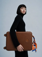 Load image into Gallery viewer, A model in a black ribbed turtleneck sweater holds a brown leather suitcase with a matching circular logo badge. The suitcase has a small orange and blue patterned scarf tied to its handle.
