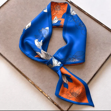 Load image into Gallery viewer, A silk scarf with a vibrant blue and orange floral pattern, styled in a knot on a neutral beige textured tray.
