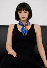 Load image into Gallery viewer, A woman with a short dark bob sits on a black leather couch wearing a black sleeveless top and a blue floral neck scarf with red and orange accents.
