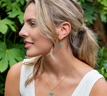 Load image into Gallery viewer, A woman in a white ribbed top wearing a delicate gold necklace and matching gold hoop earrings with small turquoise blue flower-like charms, set against a lush green outdoor background.
