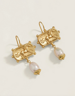 A pair of gold drop earrings featuring an embossed horse motif in an oval frame, with a dangling natural pearl in a gold dome setting, photographed on a light cream background.