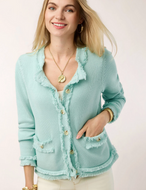 A woman wearing a light mint green fringed cardigan with gold buttons, styled with a white top and cream pants against a plain light background.