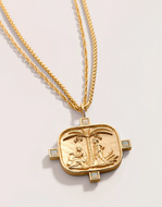 A gold pendant necklace featuring an embossed religious scene on a rounded rectangular medallion, suspended from a braided chain on a light gray background.