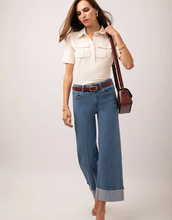 Load image into Gallery viewer, A woman modeling a cream short-sleeve button-up shirt, wide-leg blue jeans with rolled cuffs, a brown belt, and a dark brown crossbody bag against a light gray studio background.
