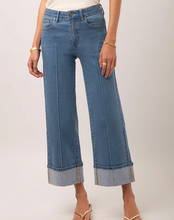 Load image into Gallery viewer, A model wearing a pair of wide-leg cropped blue denim jeans with rolled cuffs, styled with a white top and gold sandals against a light gray studio background.
