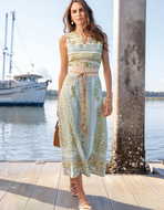 A woman stands on a wooden dock by the water wearing a sleeveless midi dress with a light blue base and green botanical pattern, a beige waist tie, and gold-toned sandals. A boat and concrete pier columns are visible in the background.