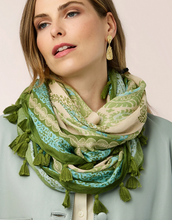 Load image into Gallery viewer, A close-up of a woman wearing a green and beige patterned scarf with tassels, gold statement earrings, and a light blue-gray jacket against a soft light pink background.
