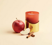 Load image into Gallery viewer, A lit candle in a translucent red glass vessel with a reflective gold base, styled with a red apple, a sliced apple piece, and two cinnamon sticks on a warm cream background.
