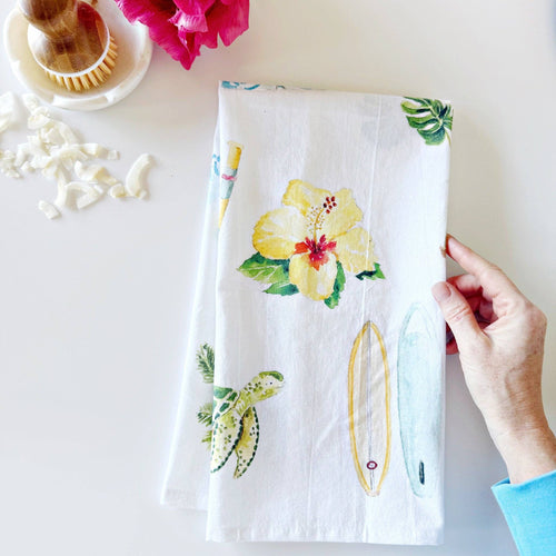 a hand holding white tea towel with tropical flower, sea turtle and surfboard designs in a watercolor style 