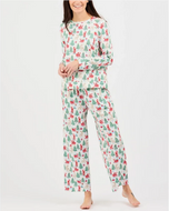 A woman modeling a matching long-sleeve top and pants pajama set with a festive Christmas print featuring pine trees, Santa Claus, and snowman motifs on a white background.